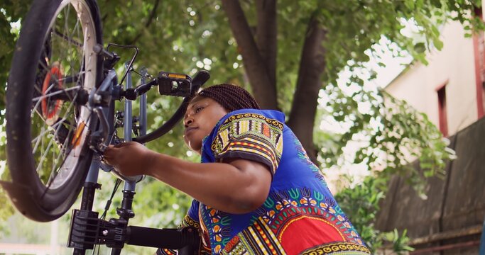 Young sports-loving female engrossed in fixing modern bicycle with professional equipment. Active determined black woman repairing bike rim and inspecting tire for outdoor leisure cycling. - Powered by Adobe