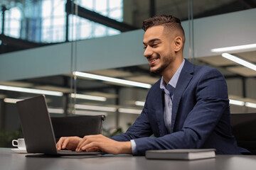 A cheerful young middle eastern businessman sits at a sleek worktable in a modern office.