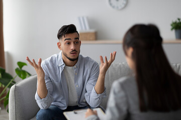 In a bright counselor's office, a young arab man sits on a couch, visibly emotional during a conversation with his psychologist. He seeks professional help for his mental health struggles.
