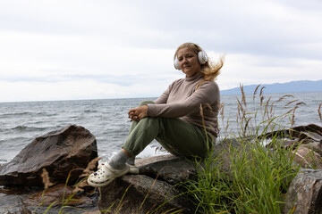 Fototapeta premium A white woman with long blonde hair listens to music with headphones, relaxing on the shore of Lake Baikal.