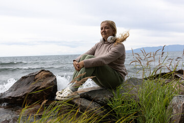 A white woman with long blonde hair listens to music with headphones, relaxing on the shore of Lake Baikal.