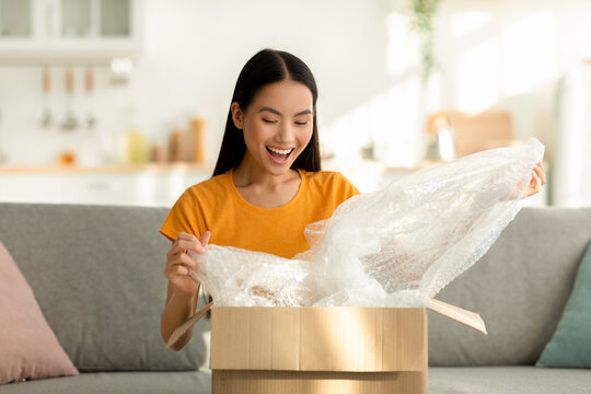 The asian woman sits on her sofa, smiling with joy as she unpacks a cardboard box filled with her online purchase. Her excitement fills the cozy living room, showcasing a moment of delight.