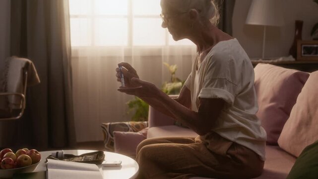 Medium backlit shot of elderly Caucasian woman with diabetes sitting on couch while testing blood sugar level, stabbing finger, dabbing blood on strip and inserting into glucose meter