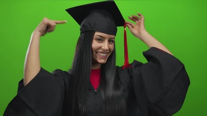 Young hispanic woman in graduation uniform smiling joyfully against isolated green background adjusting cap confidently signaling achievement and pride in her accomplishment