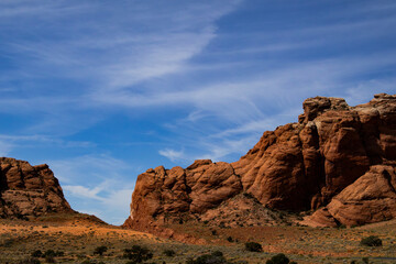 Fototapeta premium Dramatic red sandstone cliffs towering over a desert landscape under a wide, streaky blue sky.
