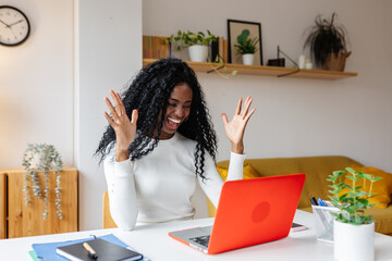 Happy young African American businesswoman or student raising arms in front of laptop after receiving good news, celebrating success while working from home