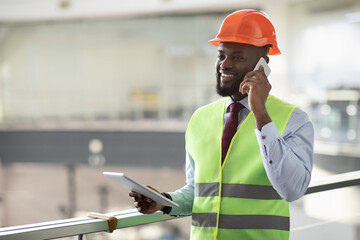 Cheerful african american young male architect in safety workwear having phone conversation with colleagues, using digital tablet, looking at copy space and smiling, side view, panorama