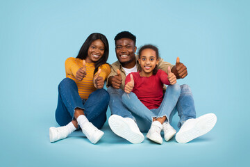 A happy family consisting of a mother, father, and daughter sits against a light blue background. They are smiling and showing thumbs up, expressing joy and togetherness in a fun, casual moment.