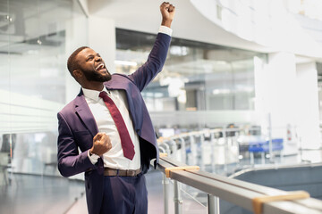 Cheerful african american young businessman celebrating success, handsome black manager in suit and tie raising hands up and screaming, office building interior, panorama with copy space