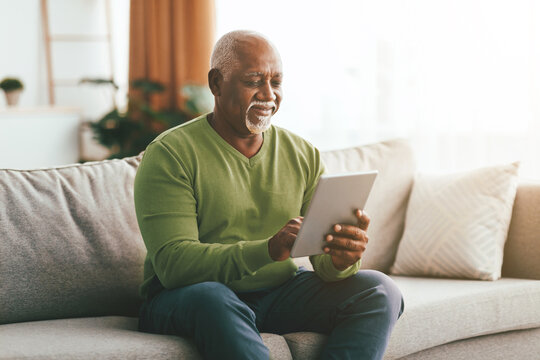 An older man sits on a soft sofa, happily engaged with a tablet in a well-lit living room. Sunlight streams through large windows, creating a warm atmosphere.