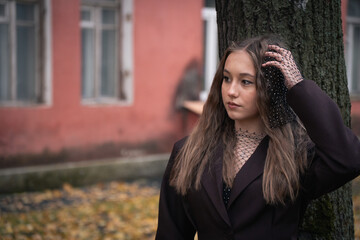 Fototapeta premium Young woman in a black mesh top and dark blazer stands near a tree with a muted red building in the background, looking away thoughtfully