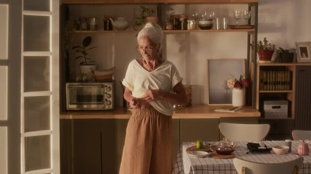 Medium full shot of elderly gray-haired Caucasian woman lifting up white blouse and injecting insulin in abdomen, after having lunch or daytime tea in kitchen at home