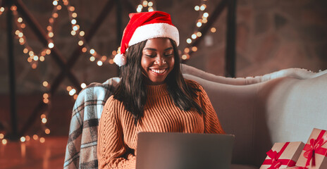 A woman wearing a red Christmas hat smiles as she shops online from her laptop. She sits comfortably on a couch surrounded by holiday decorations and gift boxes.
