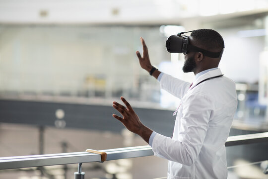 Back view of young black doctor in workwear and stethoscope using VR glasses, touching copy space, clinic interior, panorama. Modern technologies in healthcare, telemedicine concept