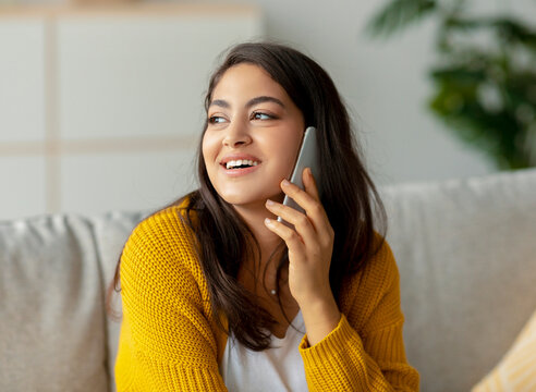 A cheerful middle eastern lady sits on her sofa in the living room, happily talking on her mobile phone. She glances aside as she enjoys her conversation with a friend.