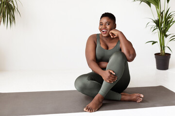 Young overweight black woman sits on a yoga mat with a big smile, enjoying her yoga practice at home in the morning. Bright and peaceful atmosphere enhances her joyful expression.