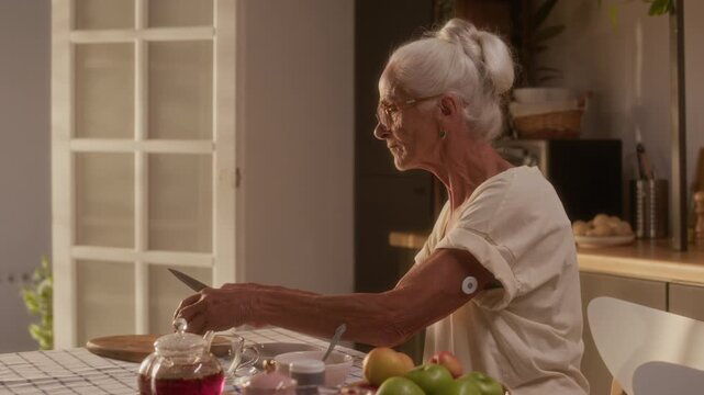 Medium shot of senior Caucasian woman with diabetic patch on arm, wearing short sleeve blouse, sitting at kitchen table, cutting up apple while snacking on fruit and enjoying tea at home