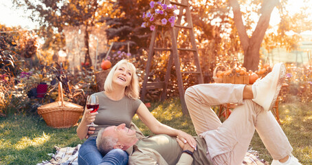 In a cozy garden filled with fall colors, a man lies on the grass while a woman sits beside him, both laughing and enjoying a glass of wine. The scene is warm and inviting.