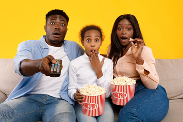 Excited young family sits on a couch in a bright yellow room, watching a movie. The parents and daughter share a moment of surprise, covering their mouths and holding popcorn.