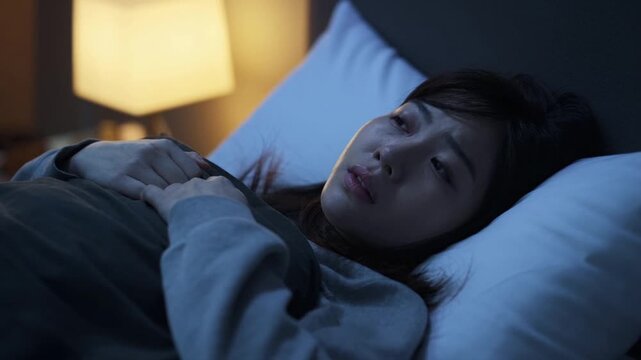Adult woman lying awake in bed at night, staring blankly with hands on blanket, apathetic mood and low energy, illuminated by soft bedside lamp, mental fatigue, stress and burnout concept