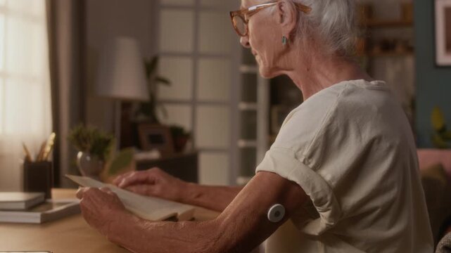 Medium close-up of elderly retired woman with autoimmune diabetes resting at table at home, reading book, touching continuous blood glucose monitor on arm and adjusting it for comfort