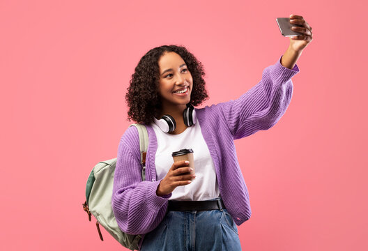A cheerful African American female, wearing headphones and a purple sweater, enjoys a takeaway coffee while taking a selfie against a pink studio background.