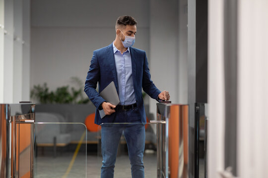 A Muslim man in a face mask stands at an electronic entrance gate, holding a laptop and access card. He is entering a modern office space during working hours, showcasing security measures in place.