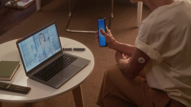 Medium shot of unrecognizable senior female patient with autoimmune diabetes and hyperglycemia learning to use CGM patch system with smartphone app, with online telemedicine consultation with GP