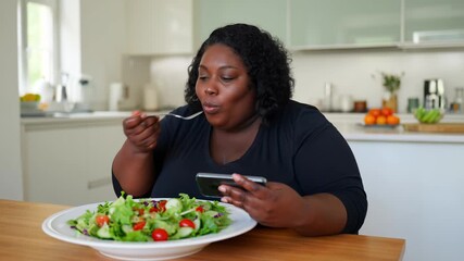 Woman eating fresh salad for breakfast at home while checking social media on a smartphone, sitting at the kitchen table with a large bowl of greens and cherry tomatoes, healthy lifestyle morning - Powered by Adobe