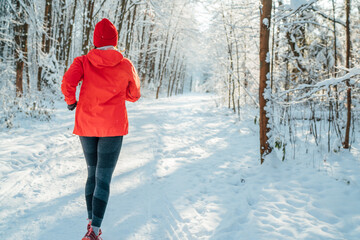Trail runner woman dressed bright red windproof jacket jogging picturesque snowy forest path during sunny frosty day. Sporty active people and winter training concept image.