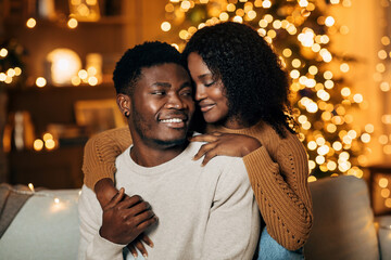 Young black couple cuddles on a couch, embracing and celebrating together on Christmas night. They enjoy a romantic moment in a beautifully decorated room filled with holiday cheer.