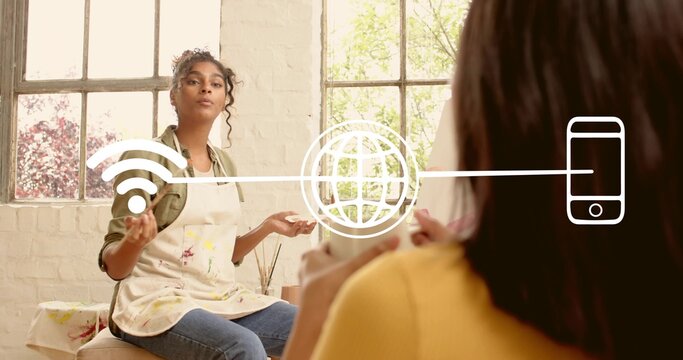 Gesturing woman artist wearing painted apron talking in bright studio with jars, mug, overlay icons