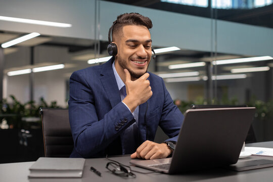 A middle eastern businessman smiles while attending an online business conference from his office. He uses a headset and sits in front of a laptop, engaging with his partners during the pandemic. - Powered by Adobe