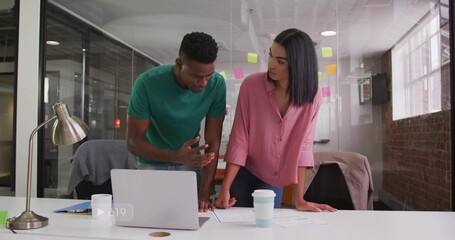 Leaning woman in pink blouse and man in green t-shirt reviewing laptop and documents at office
