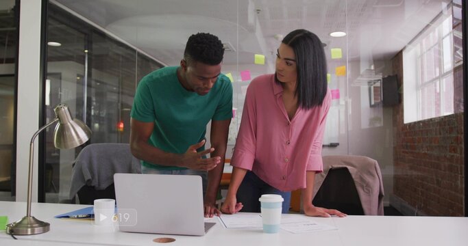Leaning woman in pink blouse and man in green t-shirt reviewing laptop and documents at office