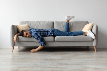 Fototapeta premium A young Middle Eastern male lies face down on a gray couch, clearly tired and drained after a long day of hard work. He is at home, looking for some rest and relaxation.