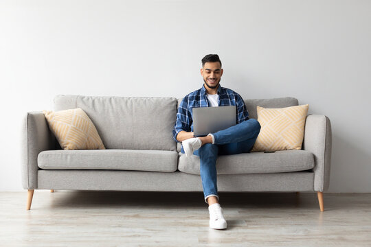 Smiling Arab man sits comfortably on a sofa at home, using his laptop. He is browsing the internet and looks cheerful while relaxing in a modern living room.