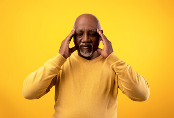 Senior African American man is touching his temples while experiencing discomfort. The orange background adds a striking contrast to his expressions of stress and discomfort related to a headache.