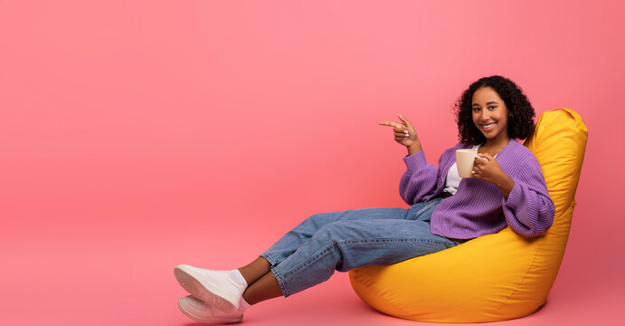 A millennial black woman relaxes in a bean bag chair while enjoying her coffee. She points to a blank space next to her, creating a cheerful atmosphere in a vibrant pink studio setting.