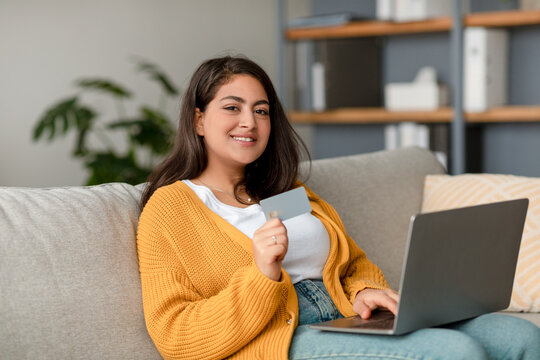 Cheerful arab woman sits on a comfortable sofa, using a laptop and a credit card to shop online. She looks content as she navigates her web banking service in a cozy home setting.
