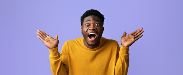 A young man wearing a yellow sweater shows excitement and happiness with open arms and a big smile. The pastel purple backdrop adds a vibrant touch to his cheerful expression.