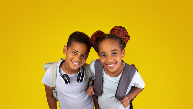 Two joyful children stand close together, smiling broadly. They wear casual clothes and carry backpacks, with a vibrant yellow backdrop that adds to their cheerful mood.