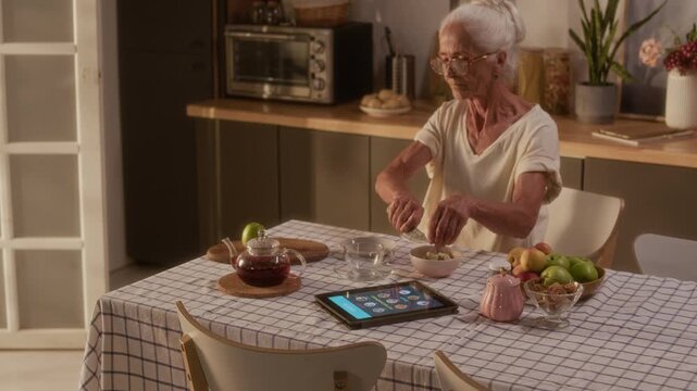 Medium shot of senior Caucasian woman, wearing blood glucose monitor patch, chopping apple and adding to oatmeal porridge for healthy breakfast in kitchen in morning, tablet with diabetic menu nearby