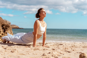 Woman in white sports clothing performing Upward Facing Dog yoga pose on bright sandy beach by ocean. She enjoys flexibility and energy in peaceful outdoor setting