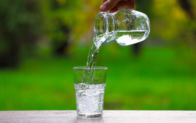 Man pouring water into glass from jug at wooden table against blurred green background, closeup. Space for text