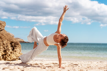 Woman in white activewear balances in Side Plank yoga pose on sunlit beach, reaching top arm toward sky. She enjoys outdoor exercise, combining strength and stability by the sea.