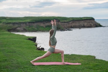Caucasian woman doing yoga on the river bank. Warrior pose.
