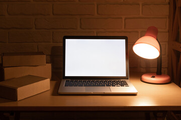 Laptop with white blank screen mockup with copy space stay on wooden desk surrounded by cardboard boxes with parcels inside, illuminated by desk lamp in evening room on cyber ​​monday or black friday.