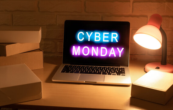 Laptop with words Cyber ​​Monday written on the screen in neon pink and blue color stands on desk surrounded by cardboard box parcels in room lit by desk lamp in the evening on the day of sale. - Powered by Adobe