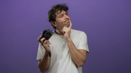 Hispanic man holding camera against purple background pondering photography techniques with thoughtful expression.